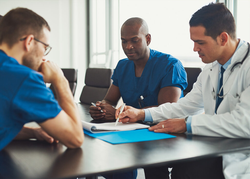 Three male doctors sit at a table and discuss items on a list.