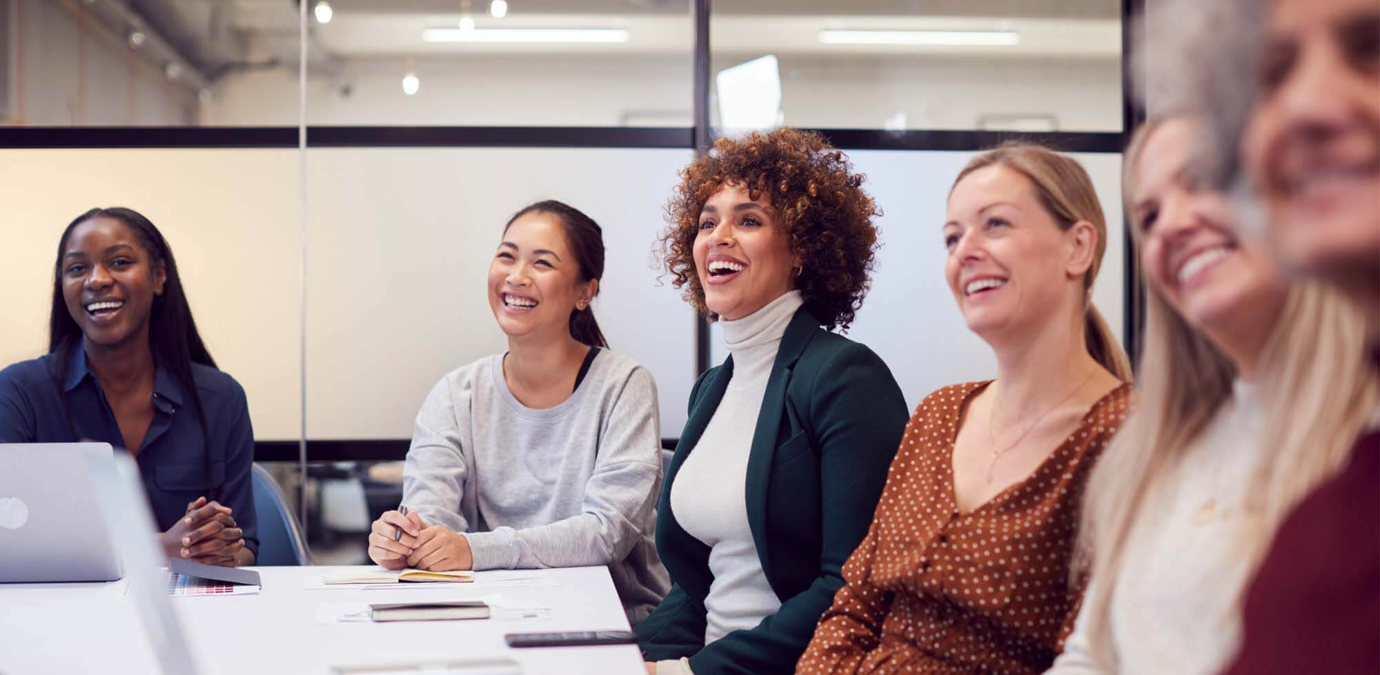 Group of women sitting around a conference room table, looking ahead and smiling.